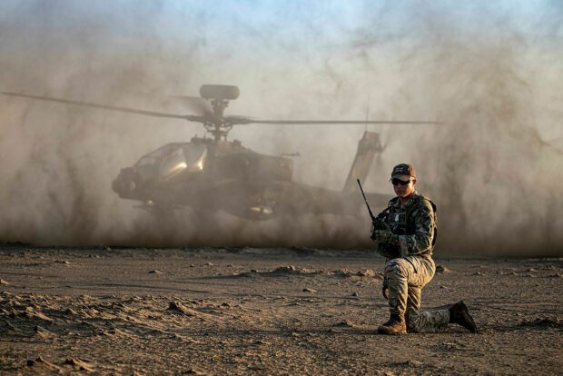 A solider kneels on dusty terrain whilst a helicopter is silhouetted in the background