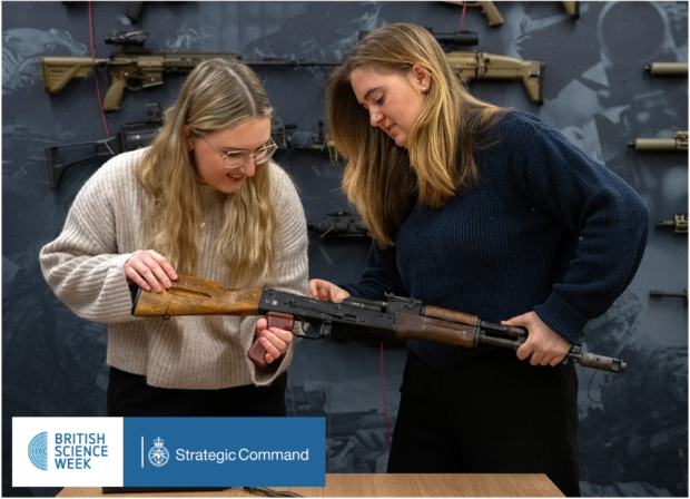 The image shows two women examining a rifle with a wooden stock. They are in a display room with various firearms mounted on the wall behind them. At the bottom of the image, there's a banner with logos for "British Science Week" and "Strategic Command"